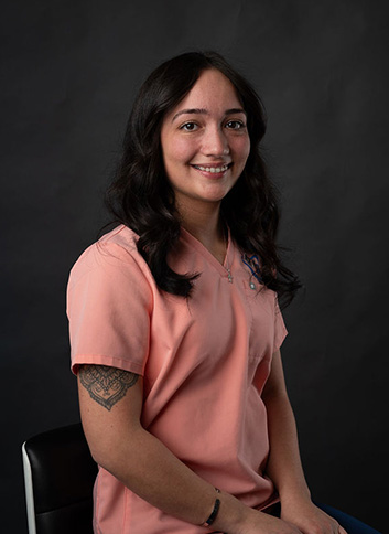 The image shows a woman wearing a light pink scrub top, sitting on a chair with a black background, smiling at the camera.