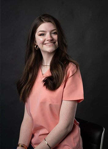 The alt text for this image could be   A young woman with long brown hair wearing a pink blouse, sitting in front of a black background.