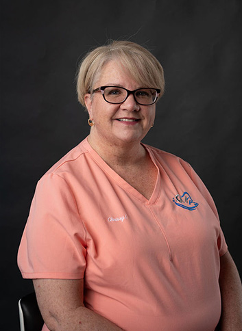 The image shows a woman wearing glasses, smiling at the camera, with short hair, sitting in a chair, dressed in a pink shirt and a white polo shirt with a logo on it.