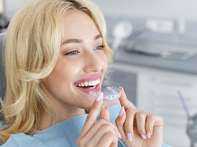 The image depicts a woman with blonde hair sitting at a dental chair, wearing a blue mask, smiling broadly while holding a small object that appears to be a toothpaste tube or dental product near her mouth.