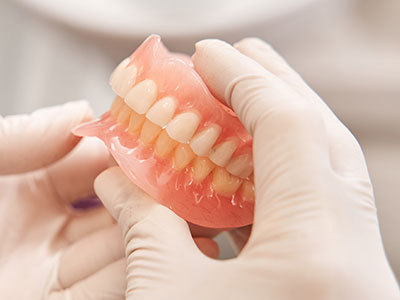 The image shows a person s hand holding a pink dental impression tray with a partial set of human teeth imprinted on it, placed in front of a pair of hands wearing white gloves, which are likely those of a dental professional examining the molds.