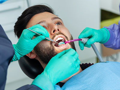 The image shows a dental professional performing a procedure on a patient s teeth while wearing protective gloves and a face mask, with another individual assisting by holding the patient s mouth open.