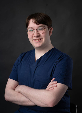 The image shows a person posing with their arms crossed, wearing glasses, a blue shirt, and white scrubs, standing against a dark background.
