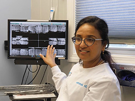 The image shows a person standing at a dental workstation with a monitor displaying X-rays, wearing a white lab coat, glasses, and a name tag.