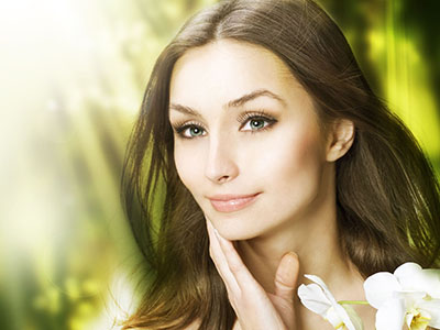 The image features a woman with fair skin and long hair, looking directly at the camera with a gentle expression. She has makeup on her face, including mascara and lipstick, and appears to be wearing a light-colored top. Her eyes are focused and she seems calm. There is a soft blur in the background that suggests an outdoor setting with natural elements like plants or flowers.