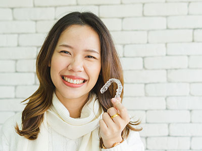 A young woman with braces smiles at the camera, displaying a toothbrush with clear bristles.